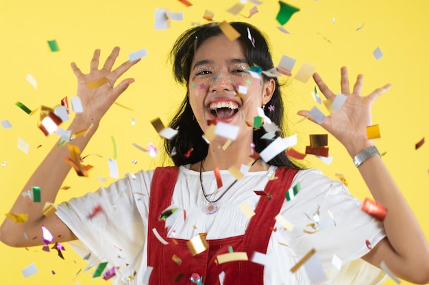 Beautiful young woman throwing confetti and looking happy on yellow background