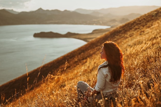 A beautiful young woman tourist is sitting on top of a mountain and enjoying the view of the sunset