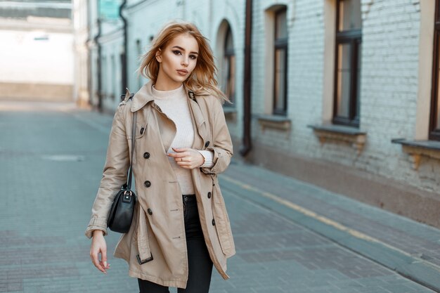 Beautiful young woman in a trendy coat with a black bag walks in the city