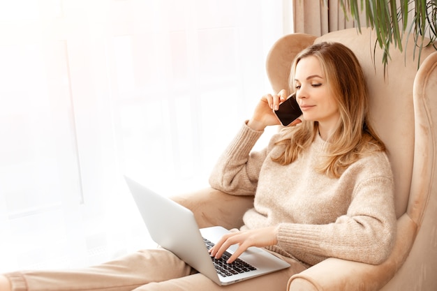 Beautiful young woman typing on laptop and call phone sitting in a chair at home near the window