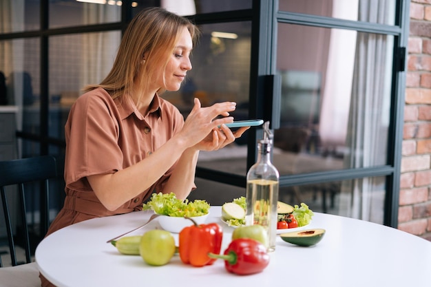 Beautiful young woman uses smartphone for taking photos of vegetables lying on kitchen table Concept of social networks Concept of healthy eating
