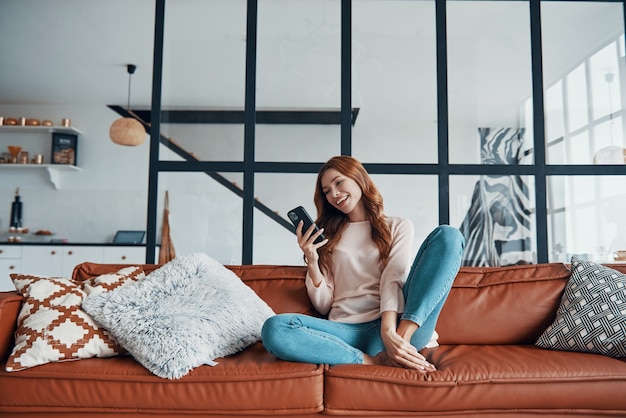 Beautiful young woman using smart phone and smiling while sitting on the sofa at home