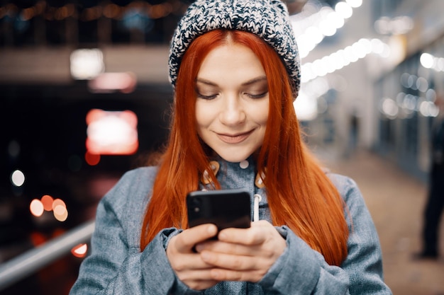Красивая молодая женщина с помощью смартфона, стоящая на улице ночного города, полной света. Портрет великолепной улыбающейся женщины с помощью мобильного телефона.