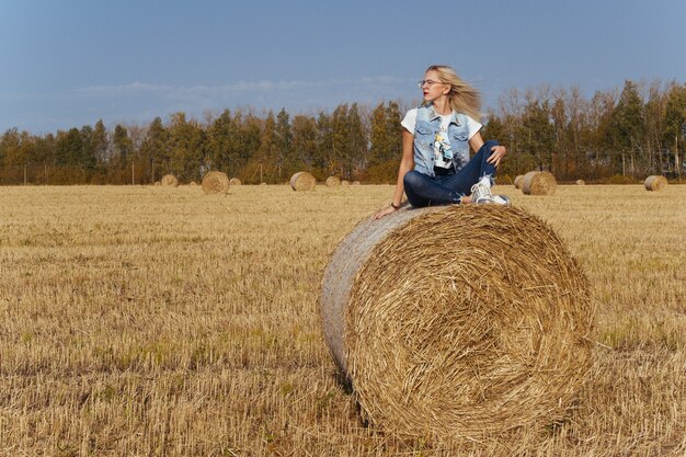 Красивая молодая женщина селян позирует в джинсах на тюке сена в поле