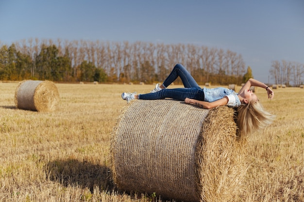 Beautiful young woman villager posing in jeans on a bale of hay in a field