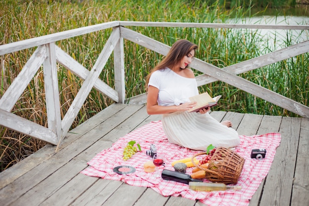 Beautiful young woman in vintage clothes has picnic on wooden pier alone. French style picnic outdoors