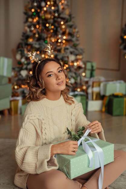 Beautiful young woman in a voluminous knitted sweater waiting for the New Year at home in the interior near the Christmas tree with a gift in her hands