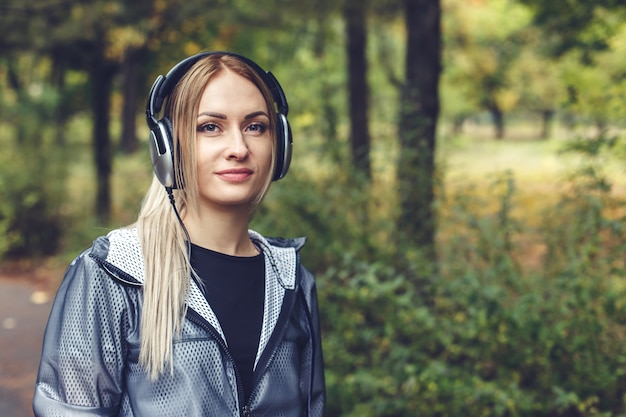 Beautiful young woman walking on city park, listening to music on headphones.