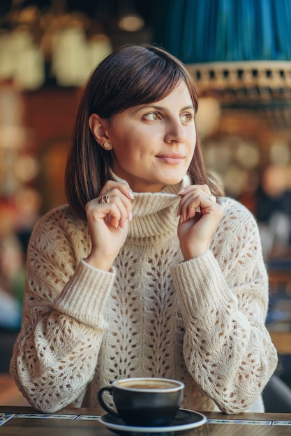 コーヒーと窓の近くのカフェで暖かいセーターを着た美しい若い女性。居心地の良い秋または冬の朝。