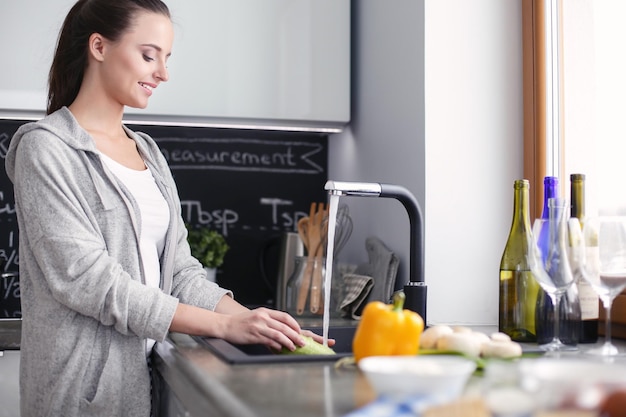 Beautiful young woman washing vegetables for salad while standing in the kitchen