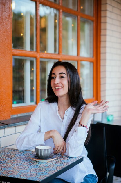 A beautiful young woman waves to passers-by at a table in a cafe. A young pretty girl wants to make an order.