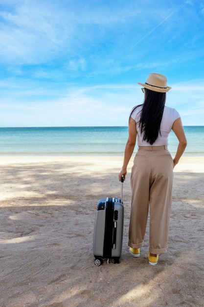 Bella giovane donna indossa t-shirt, pantaloni lunghi e cappello di paglia con una valigia su una spiaggia tropicale.