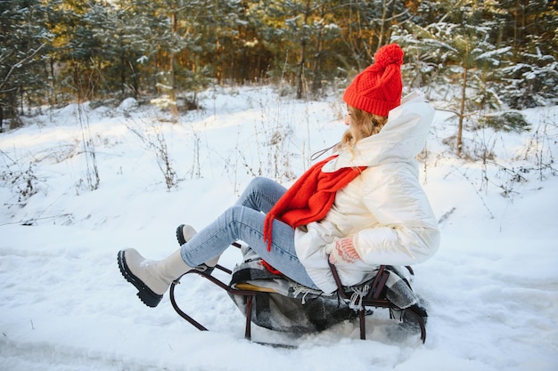 Фото Красивая молодая женщина в теплой зимней одежде сидит на санях и скатывается с холма, развлекаясь в снежный зимний день