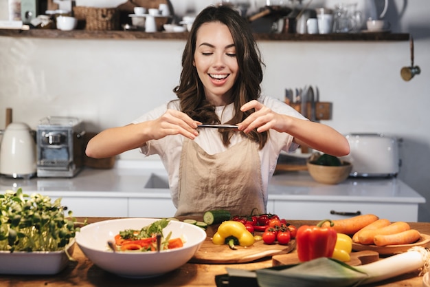 Bella giovane donna che indossa un grembiule che cucina una sana insalata in cucina a casa, scattando una foto del cibo con il cellulare