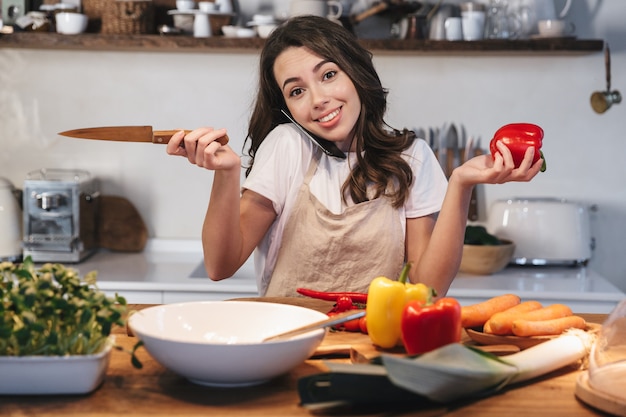 Beautiful young woman wearing apron cooking healthy salad at the kitchen at home, using mobile phone while cooking