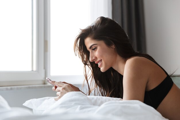 Beautiful young woman wearing black lingerie using mobile phone in bed