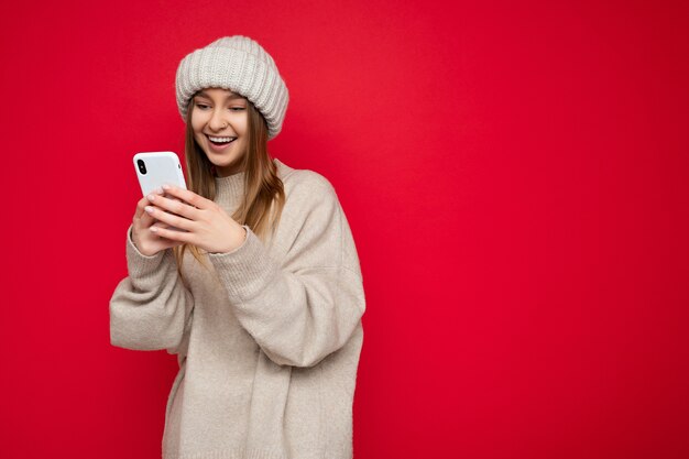携帯電話の画面を見ている電話を介してインターネット上でサーフィンを背景に孤立して立っているカジュアルな服を着ている美しい若い女性。コピースペース