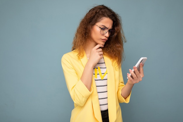 Beautiful young woman wearing casual clothes standing isolated over background surfing on the internet via phone looking at mobile screen.