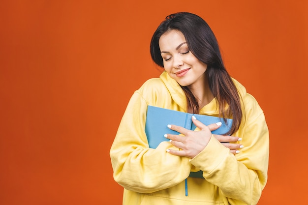 Beautiful young woman wearing casual standing isolated over orange background, reading a book.