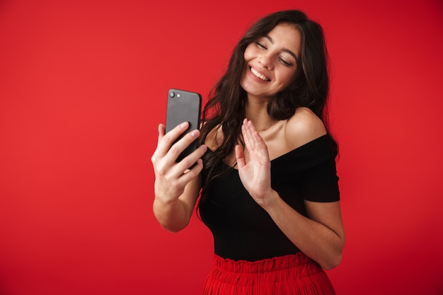 Beautiful young woman wearing a dress standing isolated over red, taking a selfie