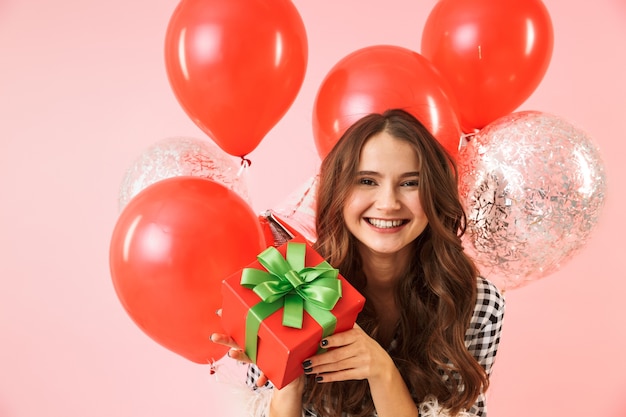 Beautiful young woman wearing a jacket standing isolated over pink background, celebrating, holding bunch of balloons and present box