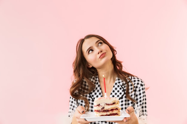 ピンクの背景の上に孤立して立っているジャケットを着て、バースデーケーキを保持している美しい若い女性