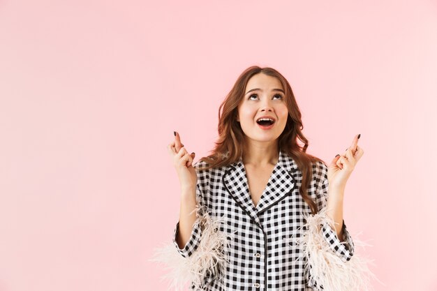 Beautiful young woman wearing a jacket standing isolated over pink background, holding fingers crossed for good luck