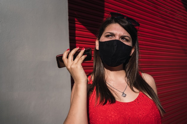 Beautiful young woman wearing a mask listening to an audio message outdoors.