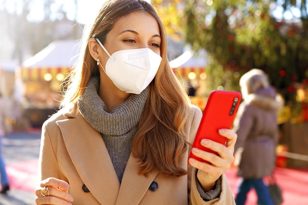 Красивая молодая женщина в медицинской маске с помощью смартфона на Рождество на открытом воздухе