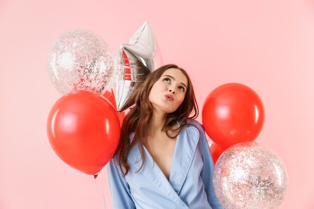 Beautiful young woman wearing pajamas standing isolated over pink background, celebrating with balloons