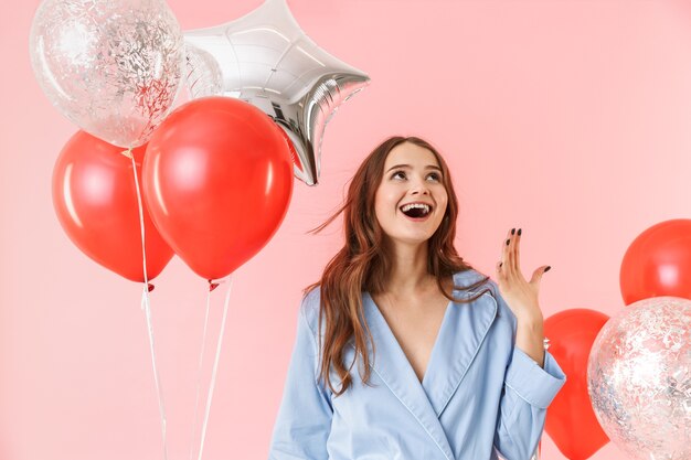 Beautiful young woman wearing pajamas standing isolated over pink background, celebrating with balloons