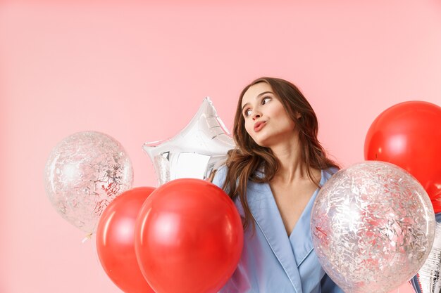 Beautiful young woman wearing pajamas standing isolated over pink background, celebrating with balloons