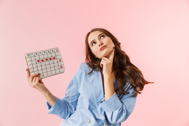 Beautiful young woman wearing pajamas standing isolated over pink background, showing menstrual calendar