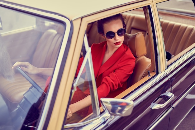 Beautiful young woman wearing red costume and sunglasses sitting in retro car