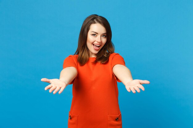 Beautiful young woman wearing red orange dress outstretched hand to camera standing posing isolated over blue wall background, studio portrait. People lifestyle fashion concept. Mock up copy space.