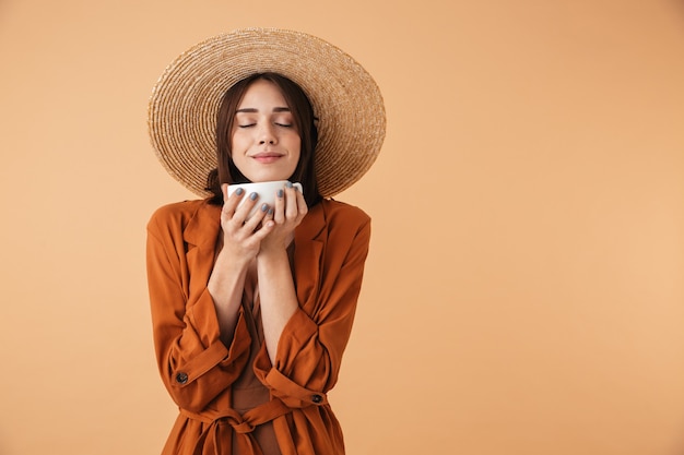 Bella giovane donna che indossa un cappello di paglia e un vestito estivo in piedi isolato su un muro beige, con in mano una tazza