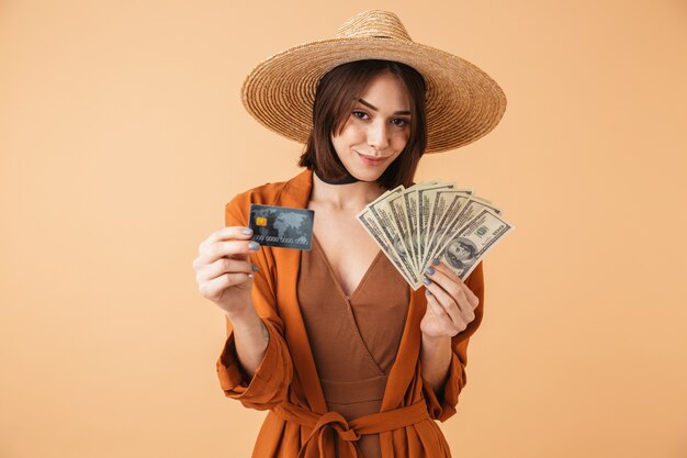 Beautiful young woman wearing straw hat and summer outfit standing isolated over beige wall, holding palstic credit card, showing money banknotes