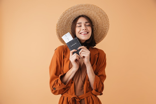 Beautiful young woman wearing straw hat and summer outfit standing isolated over beige wall, holding passport with flight tickets