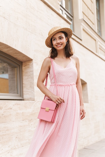 beautiful young woman wearing summer dress and straw hat smiling while walking on city street