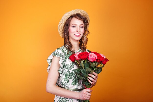 Beautiful young woman wearing summer dress and straw hat with roses bouquet on yellow background