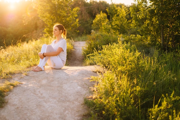 Beautiful young woman wearing white clothes sits among the green grass and trees and looks away Happy Lady resting outside in park evening on background of soft sunlight