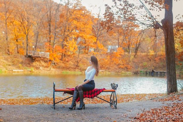 Beautiful young woman in a white blouse sits on a bench in the park in autumn. Portrait of a woman in the park with a lake and trees.