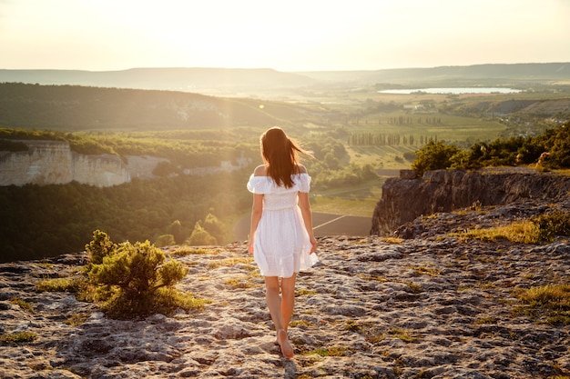 Beautiful young woman in a white dress enjoys stunning scenery in the mountains during sunset