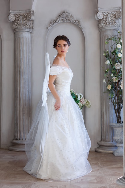 A beautiful young woman in a white dress with angel wings on her back. It stands in a beautiful decor against the wall.