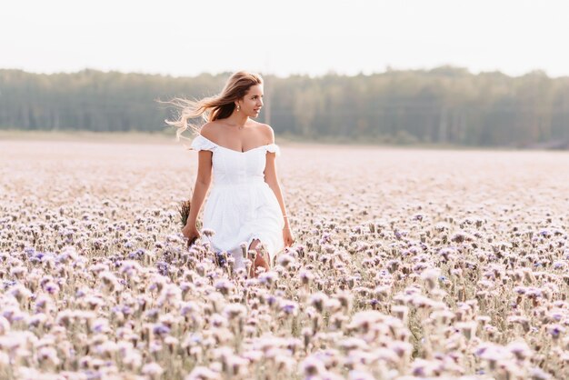 Beautiful young woman in a white dress with a bouquet of flowers in a field in summer in nature