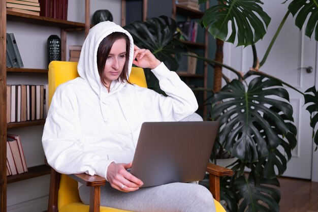 Beautiful young woman in white hoodie using laptop sitting on yellow armchair at home.