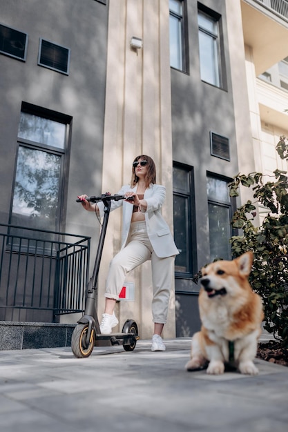 Beautiful young woman in white suit standing with her electric scooter and Corgi Dog near modern building in city and looking away