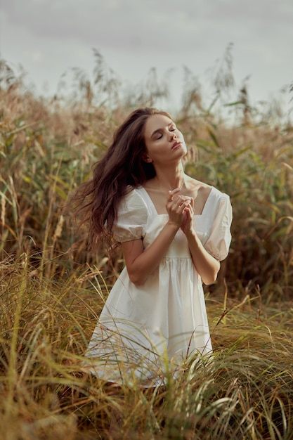 Beautiful young woman in white summer dress is sitting in tall grass in rural field Portrait of romantic girl at sunset warm sun natural beauty woman