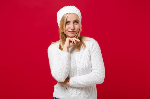 Photo beautiful young woman in white sweater, hat isolated on red background, studio portrait. healthy fashion lifestyle, people emotions, cold season concept. mock up copy space. put hand prop up on chin.