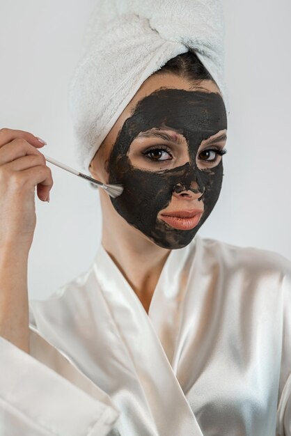 beautiful young woman in white towel applying mud black clay mask and hold brush isolated on white